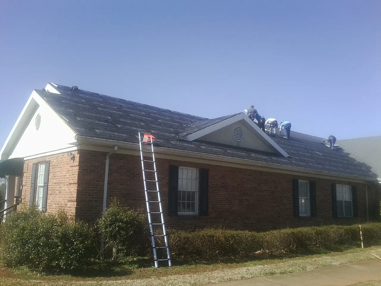 Crew tearing shingles off a brick ranch roof with a ladder set — Emergency Roof Repair work by our crew in Patchogue