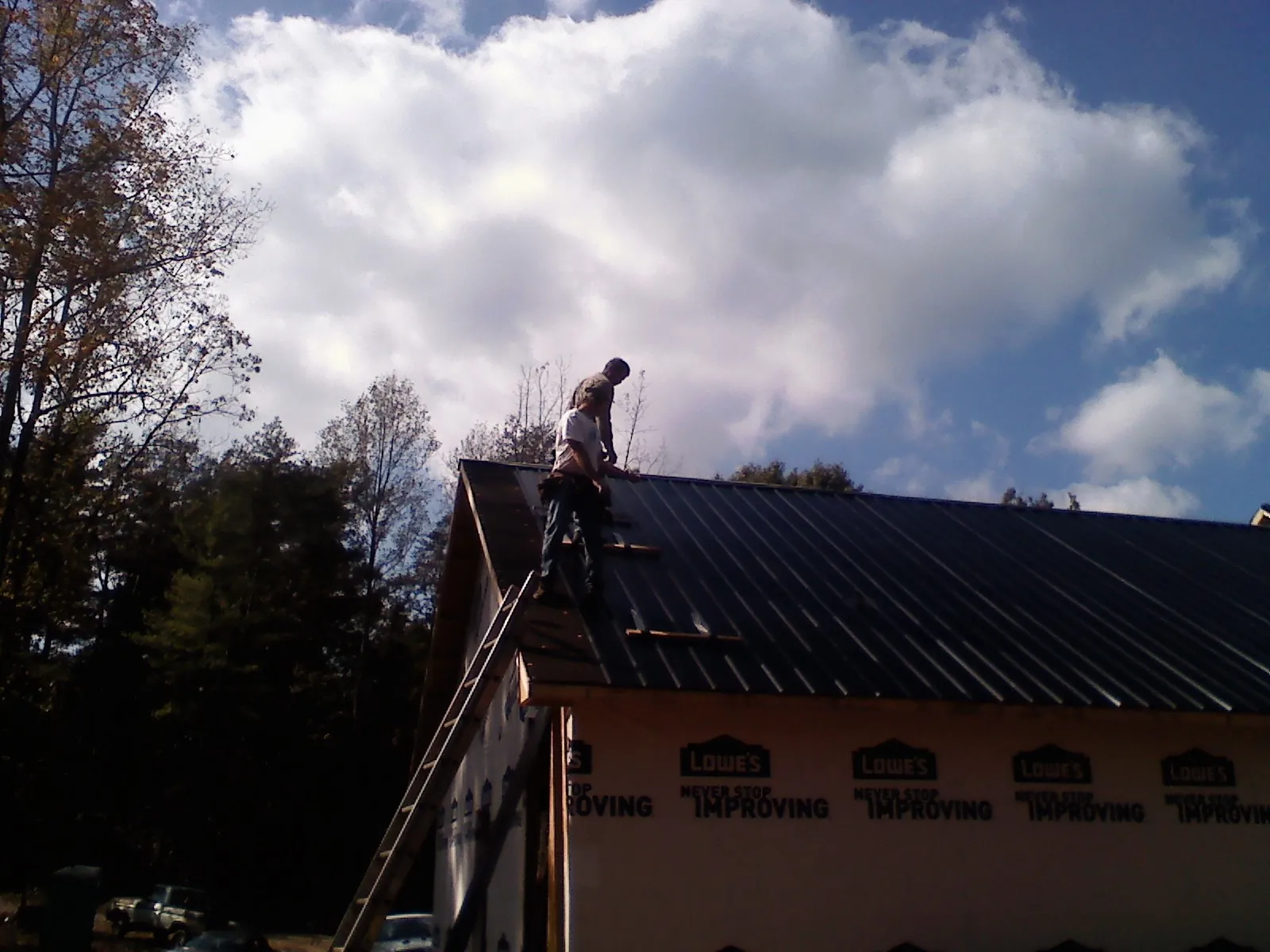 Roofer fastening dark metal panels high against a cloudy sky, from a Roof Inspection job in Patchogue